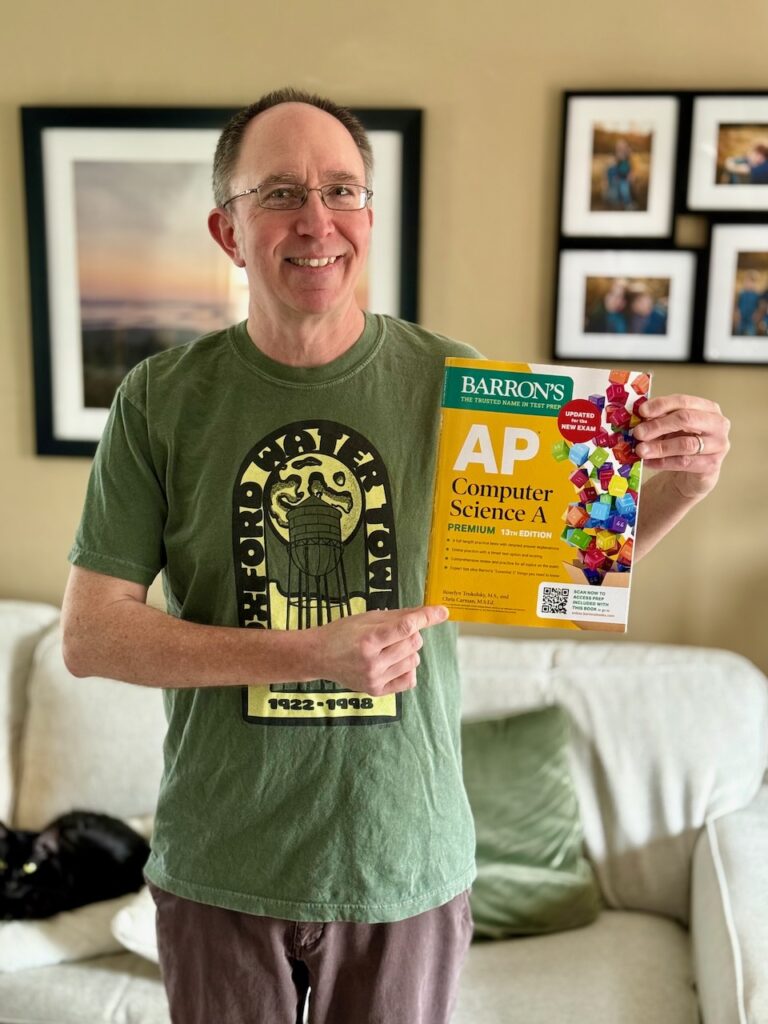 A photo of Chris Carman, a 51-year old white man wearing a green t-shirt and holding a yellow book titled "Barron's AP Computer Science A 13th Edition"