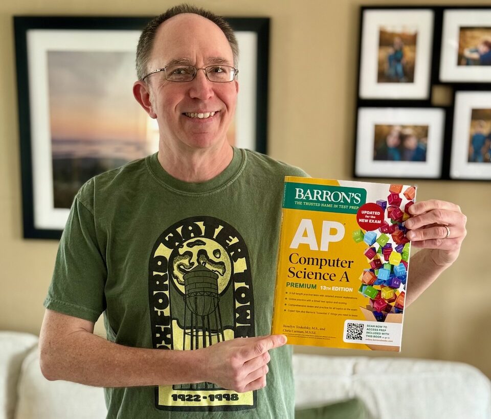 A photo of Chris Carman, a 51-year old white man wearing a green t-shirt and holding a yellow book titled "Barron's AP Computer Science A 13th Edition"