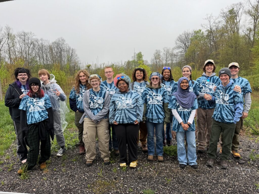 A photo shows 15 high school students standing outdoors wearing blue tie-dye t-shirts with the Roosevelt High School "R" logo on the front in white